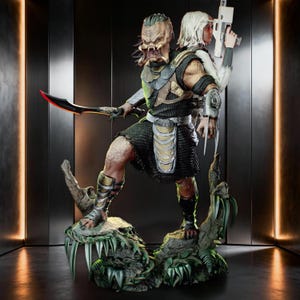 May include: A detailed statue of a Predator-like alien warrior holding a bladed weapon, paired with a human figure wielding a futuristic gun. The figures are posed on a detailed base with a jungle-like theme, featuring sharp teeth and foliage. The statue is predominantly green, brown, and silver.