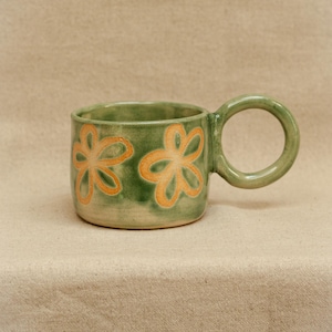 May include: A ceramic mug with a glossy sage green glaze featuring two hand-painted orange flower motifs on the exterior. The mug has a wide, cylindrical body and a large, circular handle extending from the side, showcasing a smooth finish.