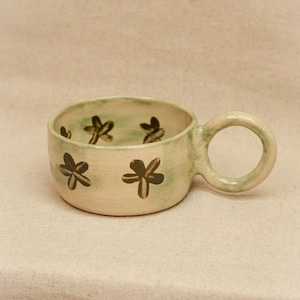 May include: A low-profile ceramic mug with a wide, circular handle. The exterior and interior feature a pale, light green wash with dark brown, hand-painted flower motifs scattered across the surface. The mug has a smooth, rounded rim.