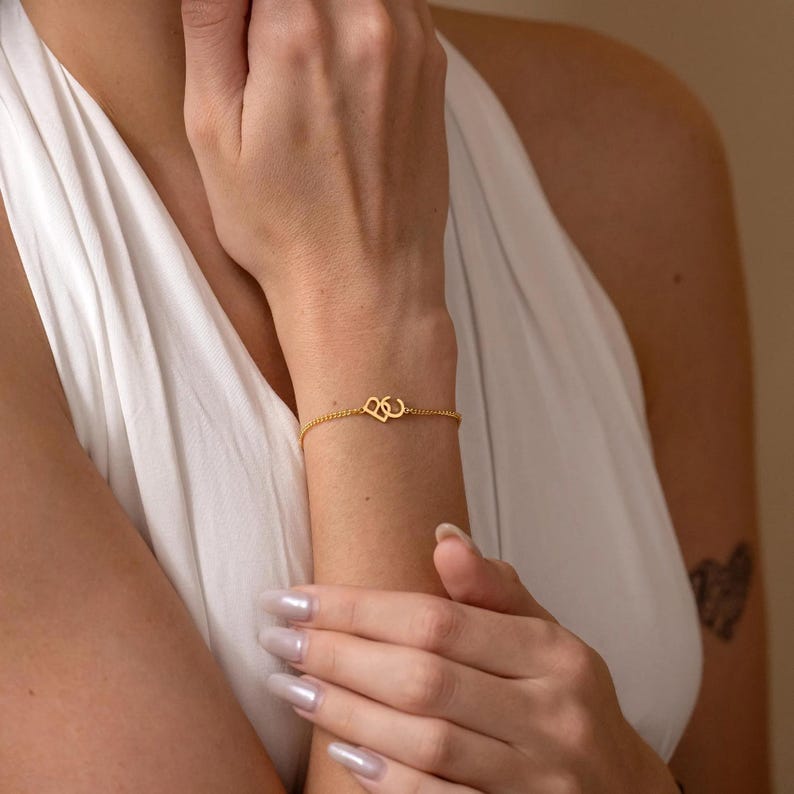 Puede incluir: Una delicada pulsera dorada con un dise&ntilde;o de doble coraz&oacute;n. La pulsera se lleva en la mu&ntilde;eca, con la cadena y los dijes de coraz&oacute;n visibles. Una joya sencilla pero elegante.
