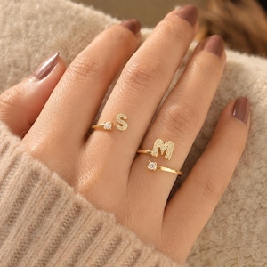 Op de afbeelding: Twee goudkleurige open ringen, elk voorzien van een letter en een kleine heldere steen. De ene ring toont de letter S en de andere ring toont de letter M. De letters hebben een textuurafwerking en de ringbanden zijn dun en gepolijst.