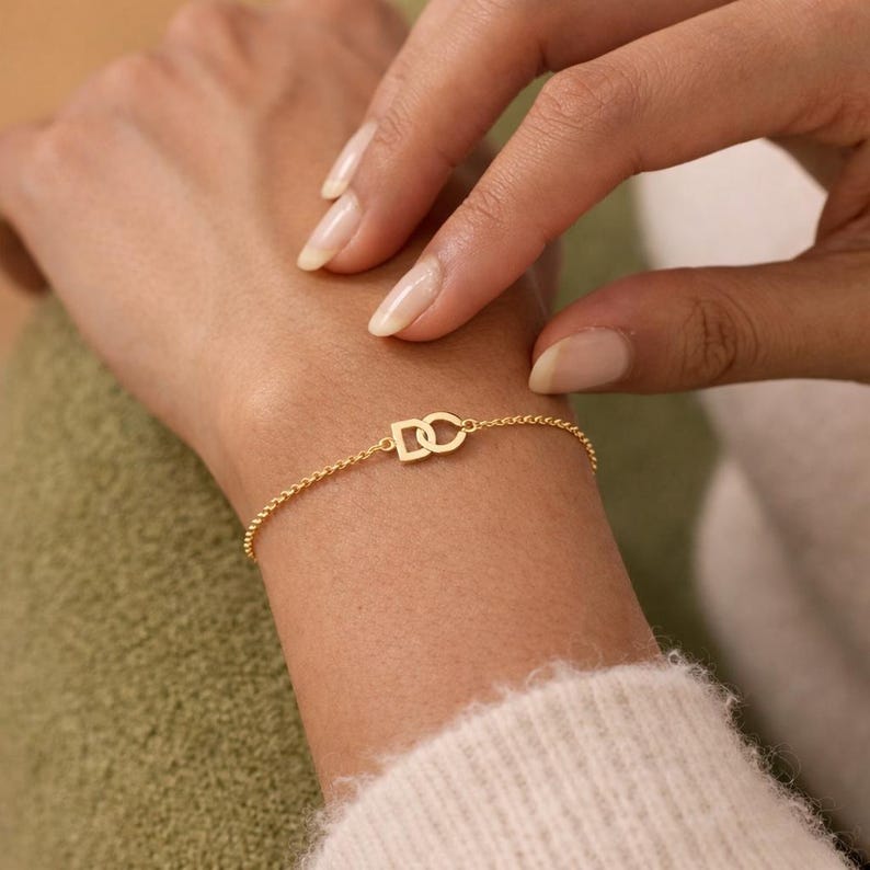 Puede incluir: Una delicada pulsera dorada con las letras entrelazadas "DC". La pulsera es una fina cadena con un peque&ntilde;o dije. La pulsera se lleva en la mu&ntilde;eca, con una mano toc&aacute;ndola suavemente.