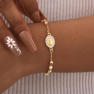 May include: A gold-toned chain bracelet featuring a central oval charm with a raised figure of the Virgin Mary surrounded by a border of small clear stones. The chain is accented with alternating small gold, silver, and rose gold-colored beads.