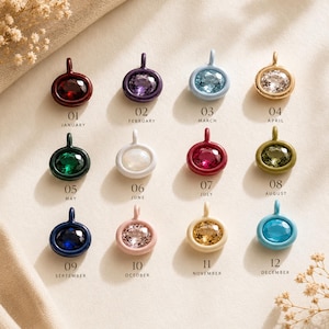 Puede incluir: Una colección de doce colgantes ovalados de piedras de nacimiento dispuestos en una cuadrícula. Cada colgante presenta una piedra preciosa de color único en un marco de metal a juego, etiquetado con un número del 01 al 12 y su mes.