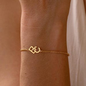 Puede incluir: Una delicada pulsera dorada con un dise&ntilde;o de coraz&oacute;n y la letra "C". La pulsera est&aacute; unida a una fina cadena, creando un aspecto minimalista y elegante. La joya se muestra en el brazo de una persona.