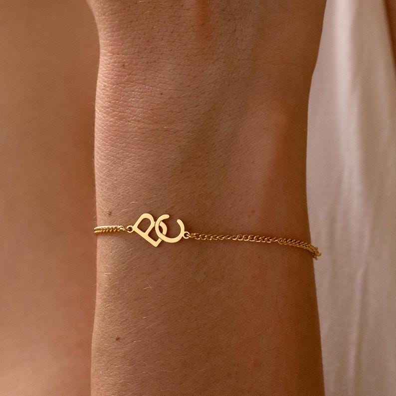 Puede incluir: Una delicada pulsera dorada con un dise&ntilde;o de coraz&oacute;n y la letra "C". La pulsera est&aacute; unida a una fina cadena, creando un aspecto minimalista y elegante. La joya se muestra en el brazo de una persona.