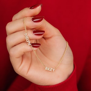 Può includere: Una collana a catena color oro con due ciondoli con nome distinti, MELYN e RILEY, ciascuno realizzato con piccole pietre chiare e scintillanti incastonate nelle lettere. La sottile catena metallica collega i due segmenti con i nomi.