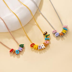 Può includere: Tre collane con sottili catene metalliche in tonalità oro e argento. Ogni collana presenta perline a forma di lettera colorate, lucide e smaltate che compongono i nomi Fiona, Charlotte e Bonnie in una disposizione curva.