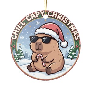 Peut inclure: Une décoration de Noël ronde en céramique représentant un capybara brun portant un bonnet de Père Noël rouge et des lunettes de soleil noires. Le capybara tient un sucre d'orge rouge et blanc. Le fond montre un paysage enneigé avec des sapins et le texte CHILL CAPY CHRISTMAS.