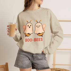 Peut inclure: Un sweat-shirt beige à manches longues orné d'un motif représentant deux abeilles déguisées en fantômes avec le texte BOO-BEES imprimé en orange en dessous. Le sweat-shirt a un col rond et est porté avec un short en jean gris.