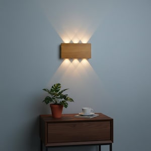 May include: A rectangular wooden wall sconce with six lights, casting beams of light. The sconce is mounted on a light blue wall. Below, a small potted plant and a cup and saucer sit on a dark wooden nightstand.