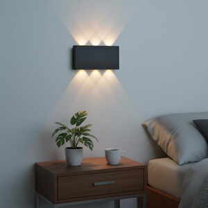 May include: A rectangular, dark gray wall sconce with three lights shining downward and three shining upward. The sconce is mounted on a light gray wall above a wooden nightstand with a potted plant and a white mug.