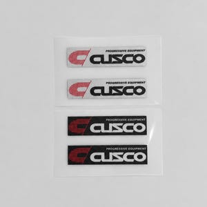 May include: Four rectangular adhesive decals featuring the Cusco logo and the text Progressive Equipment. Two decals have a silver background with black text and red accents, while two decals have a black background with white text and red accents.