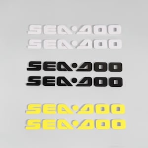 Puede incluir: Seis pegatinas dispuestas en tres pares, cada una con el texto SEA-DOO en una fuente negrita y estilizada. El par superior es blanco, el par central es negro y el par inferior es amarillo brillante. Cada pegatina tiene un diseño geométrico.