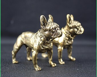 May include: Two bronze-colored French bulldog figurines. The dogs are standing, with detailed features including collars and wrinkles. The figurines have a metallic sheen and are set against a dark background.