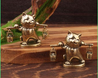 May include: Two bronze cat figurines, each carrying a yoke with two buckets. The cats have smiling faces and wear small bells. The buckets have Chinese characters. The figurines are on a wooden surface.