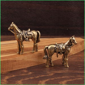 May include: Two bronze-colored horse figurines with saddles and bridles are displayed on a wooden surface. The horses are detailed with realistic features, and the overall aesthetic is vintage or antique.