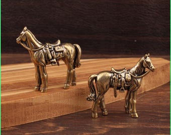 Puede incluir: Dos figuras de caballos de color bronce, con sillas de montar y bridas, sobre una superficie de madera. Los caballos presentan detalles realistas, evocando un estilo vintage o antiguo, con un tono metálico cálido.