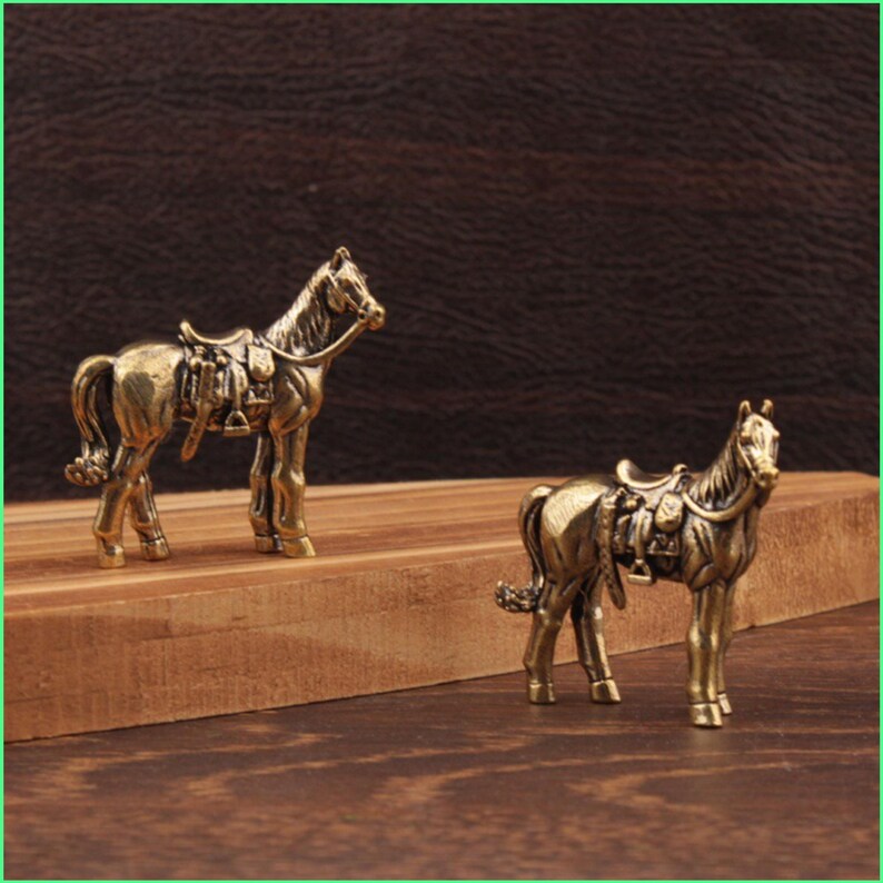 May include: Two bronze-colored horse figurines with saddles. The horses are standing on a wooden surface against a dark brown background. The figurines have detailed features, including manes, tails, and tack.