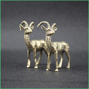 May include: Two bronze-colored goat figurines. The goats have detailed features, including curved horns, textured fur, and slender legs. They stand side-by-side against a dark background, showcasing their intricate design and craftsmanship.