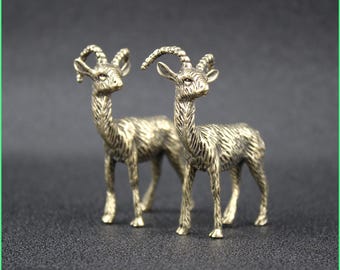 May include: Two bronze-colored goat figurines. The goats have detailed features, including curved horns, textured fur, and slender legs. They stand side-by-side against a dark background, showcasing their intricate design and craftsmanship.