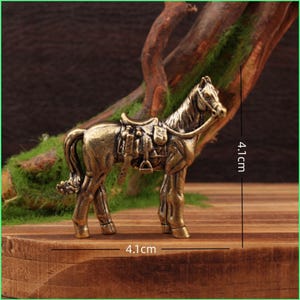 May include: A bronze-colored horse figurine, 4.1 inches long and 4.1 inches high, with a saddle and bridle. The miniature horse is standing on a wooden surface, with a backdrop of green moss and a tree branch.