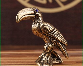 May include: A small, antique brass toucan figurine with a curved beak and a blue gem eye. The bird is perched on a textured base, showcasing detailed feather and wing patterns. The figurine has a vintage aesthetic.