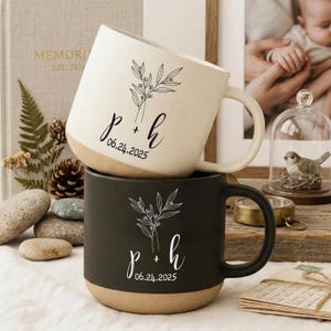 Può includere: Due tazze in ceramica, una color crema e una nera, impilate. Ogni tazza presenta un motivo floreale e le iniziali "p + h" con la data "06.24.2025" in corsivo. La base di ogni tazza è di colore marrone naturale.