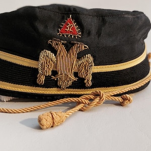 Puede incluir: Gorra negra con ribete dorado y un emblema de águila bicéfala bordado en oro. La gorra tiene un triángulo rojo con bordado dorado. Una cuerda dorada con borlas está unida a la gorra.