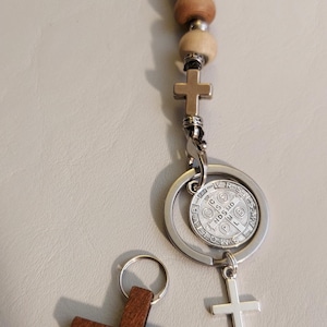 May include: A keychain featuring a strand of light brown wooden beads with silver-toned spacers, a small silver cross, and a circular Saint Benedict medal. A separate dark brown wooden cross with a metal ring is positioned next to the keychain.