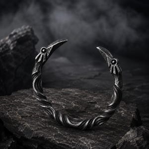 Puede incluir: Un brazalete de metal en tono plateado oscuro con una textura retorcida y en espiral a lo largo de la banda. Los dos extremos del brazalete abierto tienen forma de cabezas de ave estilizadas, cada una con una pequeña incrustación oscura.