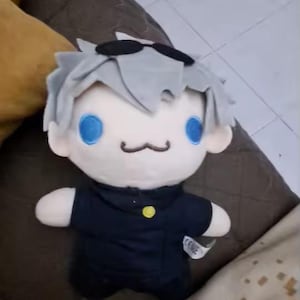 May include: A plush doll featuring spiky light gray hair, black sunglasses resting on the forehead, bright blue circular eyes, and a small brown stitched mouth. The doll wears a dark navy blue collared shirt with a single yellow button detail.