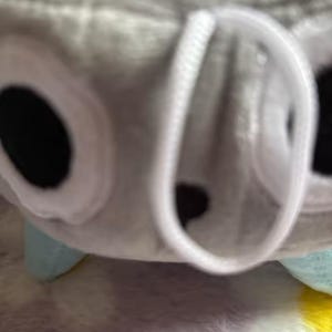 May include: A close-up of a plush toy, featuring a gray body with white accents around large black eyes. The toy has a white, circular opening and small, light blue feet. The background is a soft, textured surface.