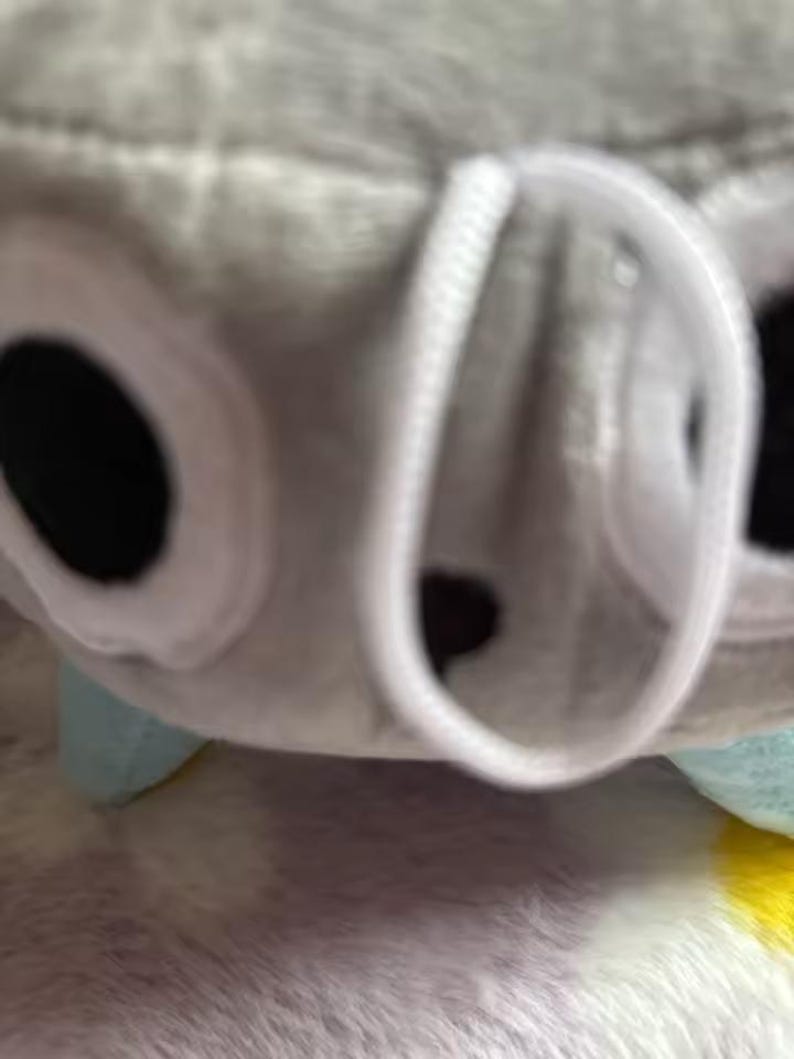 May include: A close-up of a plush toy, featuring a gray body with white accents around large black eyes. The toy has a white, circular opening and small, light blue feet. The background is a soft, textured surface.
