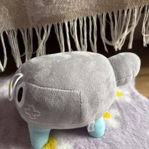 May include: A plush toy shaped like a gray, rounded creature with light blue legs and a small tail. It has a cross symbol on its side and a white tag. The toy is on a purple floral patterned rug.