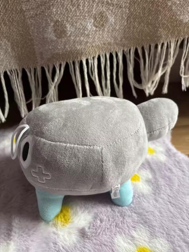May include: A plush toy shaped like a gray, rounded creature with light blue legs and a small tail. It has a cross symbol on its side and a white tag. The toy is on a purple floral patterned rug.