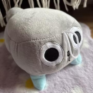 May include: A plush toy in the shape of a creature with large, round eyes and small blue feet. The toy is primarily gray with white and black accents on the eyes. The toy is sitting on a patterned surface.