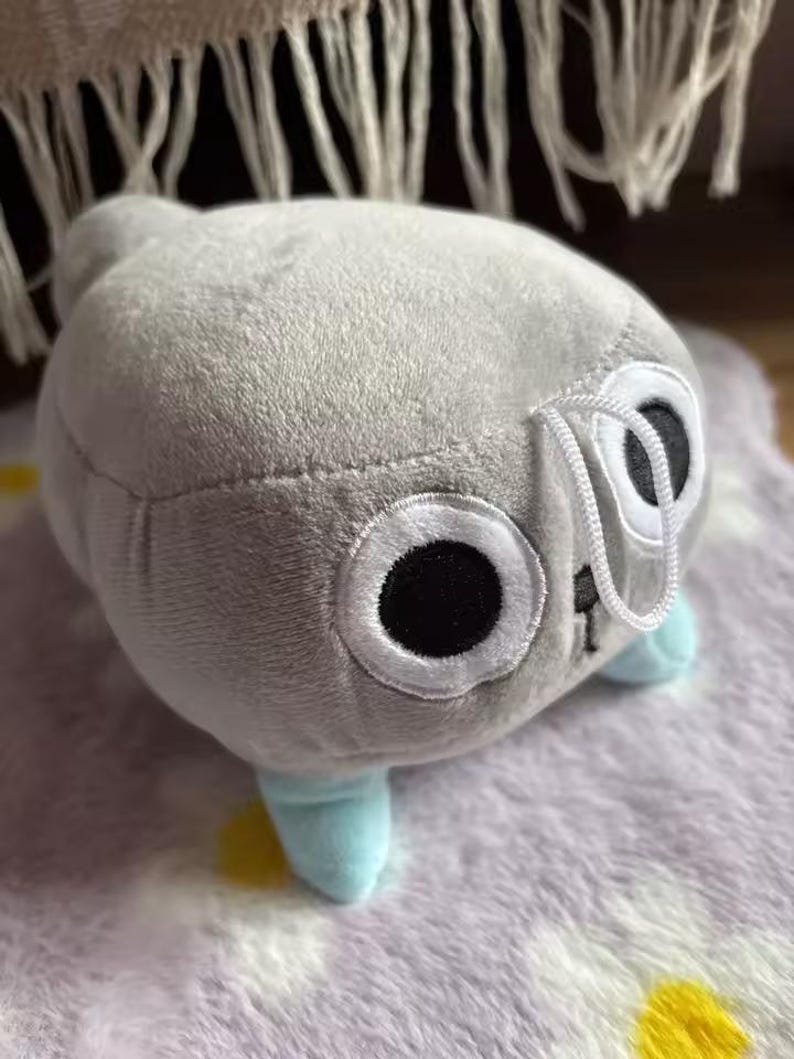 May include: A plush toy in the shape of a creature with large, round eyes and small blue feet. The toy is primarily gray with white and black accents on the eyes. The toy is sitting on a patterned surface.