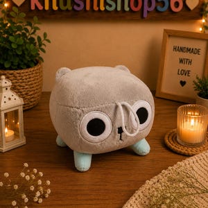 May include: A gray plush toy with large black eyes and light blue feet. The toy has a rounded body and a small white nose. The background includes a wooden sign that says "khushishop56" and a framed sign that says "HANDMADE WITH LOVE".
