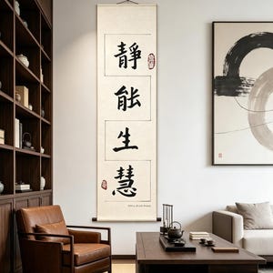 May include: A vertical scroll with Chinese calligraphy in black ink, featuring four characters within rectangular frames. The scroll is cream-colored, with a wooden rod at the bottom. A red seal is visible. The text reads "Stillness Breed Wisdom".