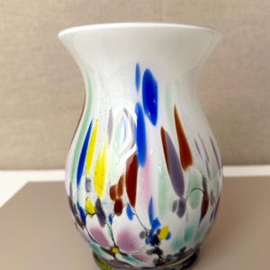 May include: A white glass vase with a flared rim and a rounded body, featuring a colorful speckled pattern of blue, yellow, purple, green, and brown elongated spots and thin dark lines throughout the exterior surface of the glass vessel.