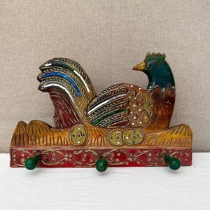 May include: A decorative wooden rooster-shaped wall hook with three green knobs. The rooster features a colorful tail, a green head, and a red and gold base. The design includes intricate patterns and details.