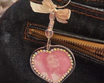 May include: A heart-shaped keychain featuring a pink-toned portrait of Michael Jackson. The charm is bordered by small iridescent rhinestones and attached to a metal ring with a cream-colored fabric bow and two clear beads.