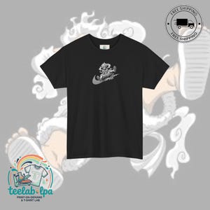 以下が含まれることがあります： 黒いTシャツ。グレーのグラフィックデザインは、スウッシュロゴの中にキャラクターが描かれています。Tシャツは半袖とクルーネックです。「teelab.lpa PRINT-ON-DEMAND & T-SHIRT LAB」の文字が下部に表示されています。「FREE SHIPPING」のロゴも表示されています。