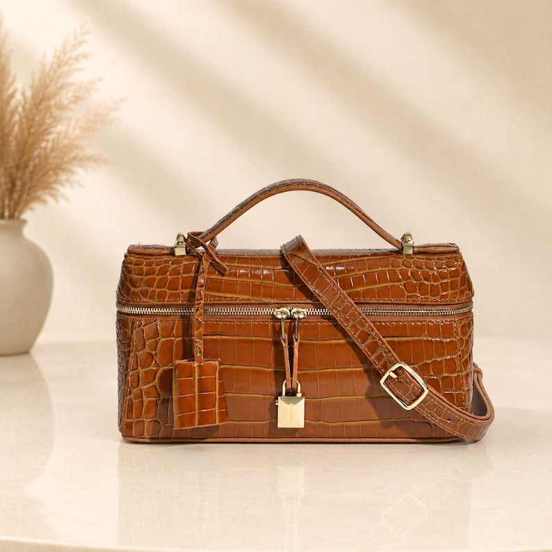 May include: A rectangular brown handbag with a glossy crocodile-embossed texture. The bag features a top handle, an adjustable shoulder strap, a gold-toned zipper closure, and a small gold padlock hanging from the center of the front panel.