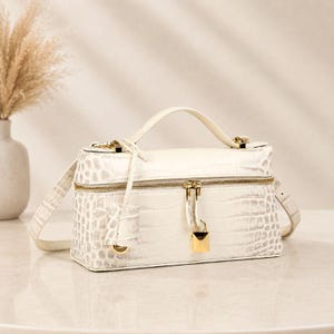 May include: A rectangular off-white handbag with a crocodile-embossed texture, featuring a top handle, a detachable shoulder strap, a gold-tone zipper closure, and a small gold-tone padlock hanging from the front zipper pull.