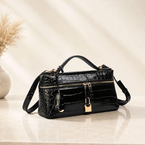 May include: A black rectangular handbag with a glossy crocodile-embossed texture. The bag features a top handle, a detachable shoulder strap, gold-tone hardware, a horizontal zipper closure, and a small gold padlock hanging from the front zipper.