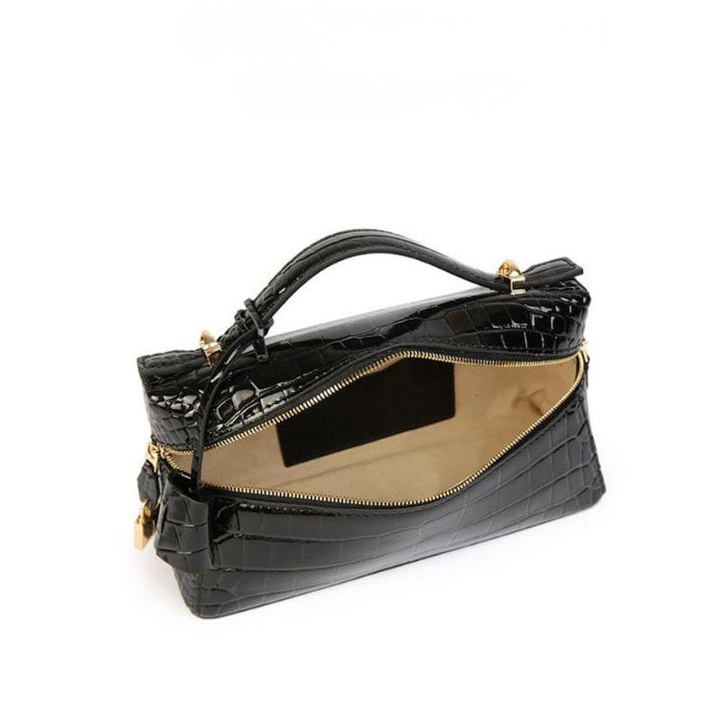 May include: A black handbag with a glossy, embossed crocodile skin texture. The bag features a structured rectangular shape, a single top handle, gold-toned metal hardware, and a wide zippered opening that reveals a light beige interior lining.
