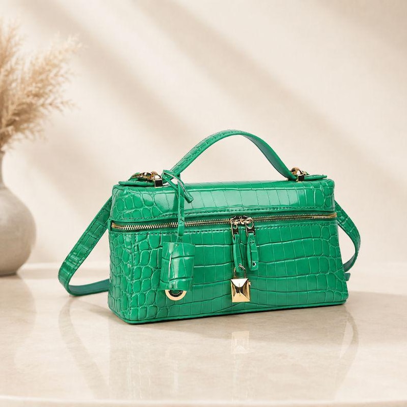 May include: A vibrant green rectangular handbag featuring a textured crocodile-embossed pattern. The bag includes a top handle, a detachable shoulder strap, a gold-toned zipper closure, and a small gold padlock charm hanging from the front zipper.