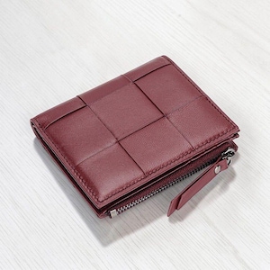 May include: A deep burgundy bifold wallet featuring a woven leather exterior pattern. The wallet includes a side compartment secured with a silver-toned metal zipper and a matching leather pull tab, showcasing a structured and compact square design.